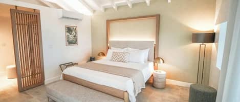 A bedroom with a bed, bedside tables, a bench, a wall-mounted air conditioner, a framed picture, and a floor lamp.