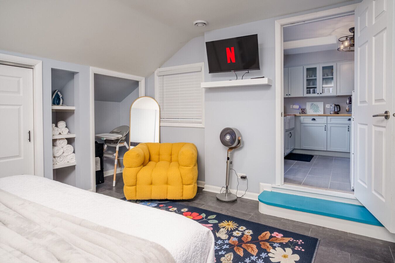 Un dormitorio con una cama, un sillón amarillo, un televisor, un ventilador y una puerta abierta hacia la cocina.