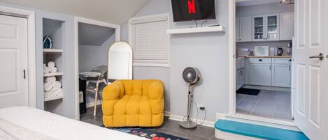 Un dormitorio con una cama, un sillón amarillo, un televisor, un ventilador y una puerta abierta hacia la cocina.