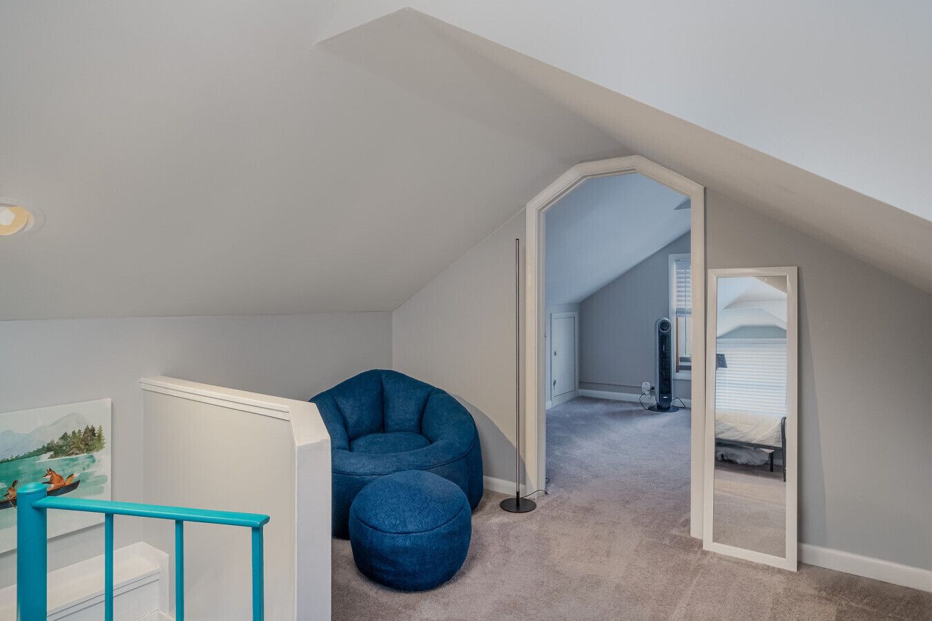 Une pièce avec un plafond en pente, un fauteuil en forme de poire de couleur bleue, un meuble blanc et un miroir qui reflète un lit et des stores.