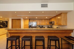 A kitchen with wooden cabinets, a stainless steel refrigerator, a microwave, and a granite countertop. There are four stools at the bar area. - Ski in Ski out Main Street Private Balcony Hot Tub Mountain Views Condo (Park City)