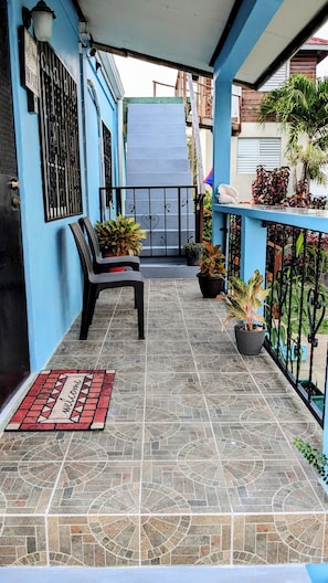 A covered balcony with a tiled floor, a bench, potted plants, and a welcome mat.