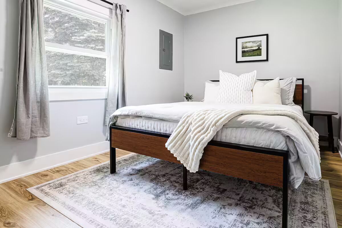 A bedroom with a bed, a window with curtains, a framed picture on the wall, and a rug on the floor.