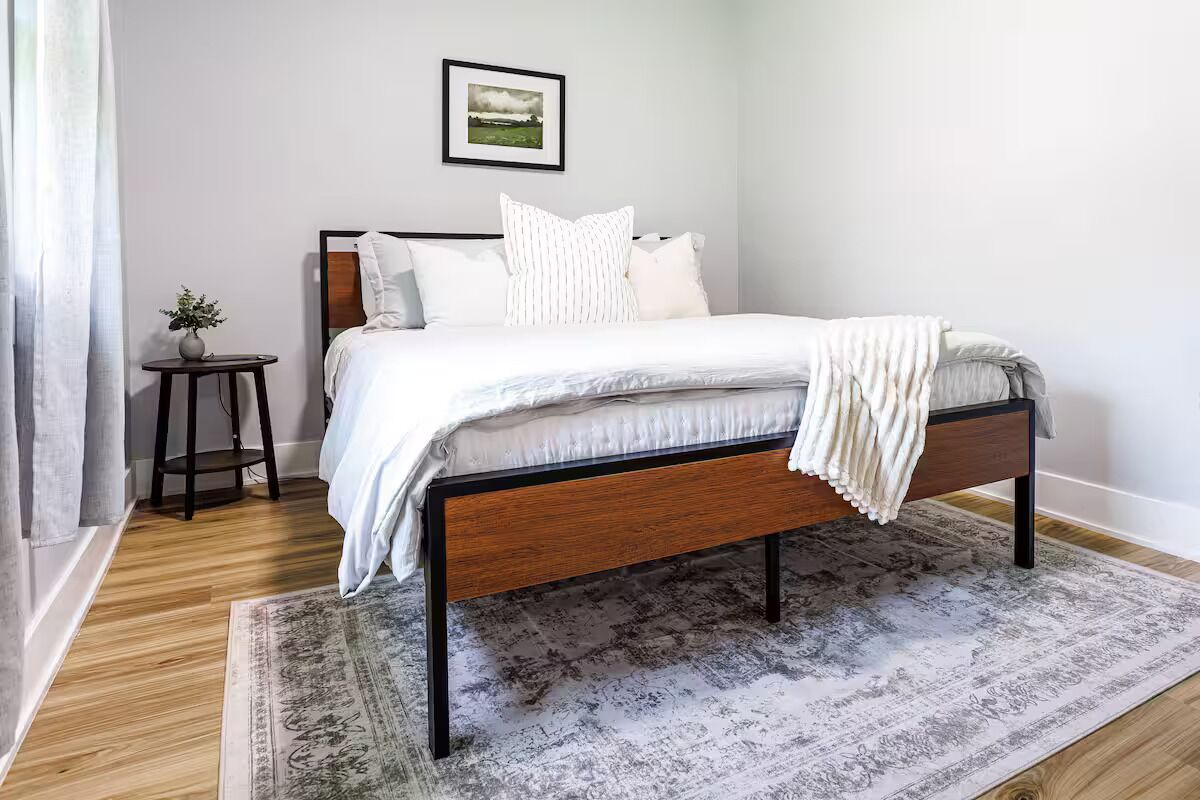 A neatly made bed with a wooden frame and white bedding, a small round table with a plant beside it, and a framed picture on the wall.