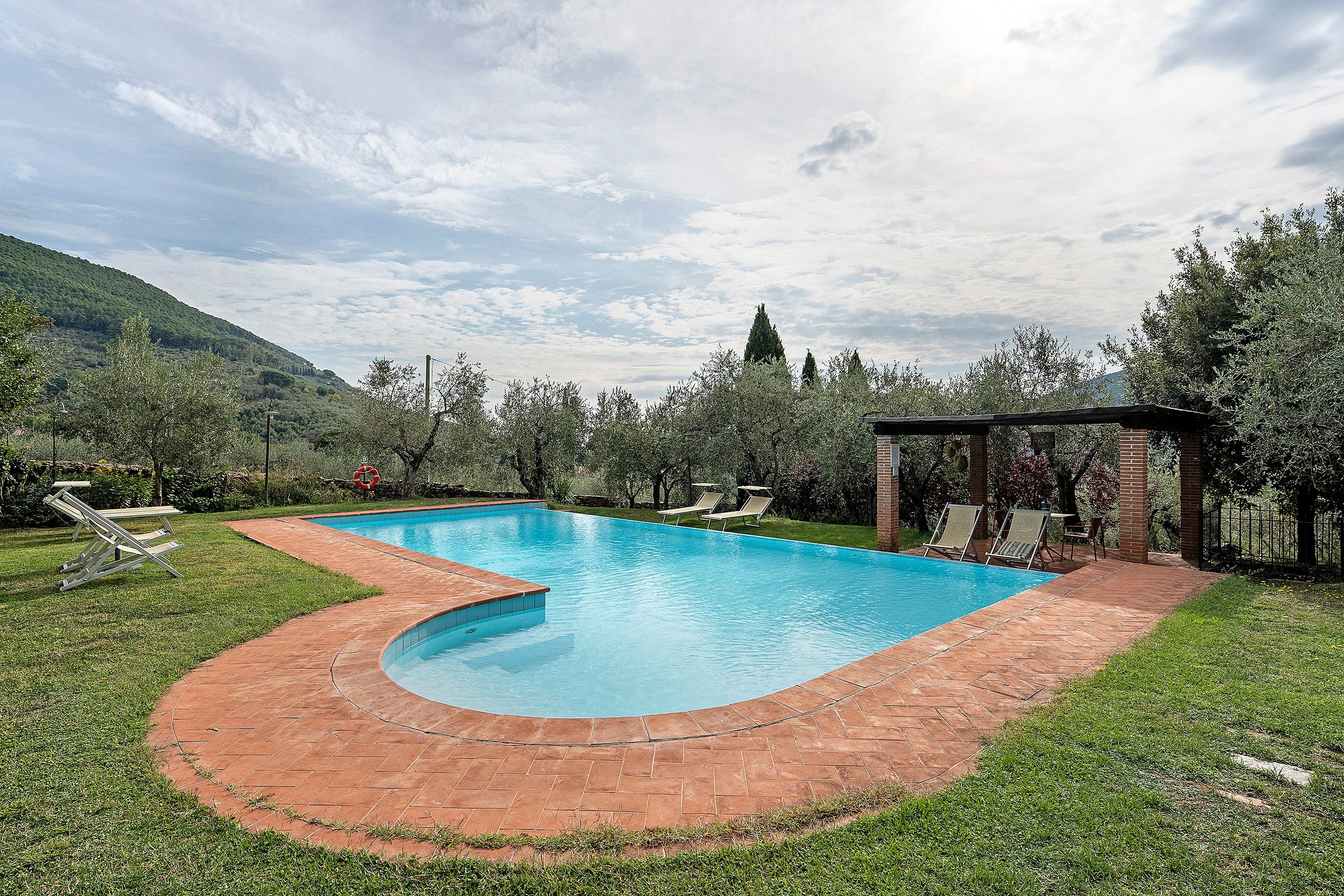 Une piscine rectangulaire à bord incurvé, entourée d’une terrasse en briques et d’une pelouse.