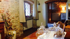 A room with a stone wall, a window with curtains, a radiator, a wooden table with glassware, and a decorative vase.