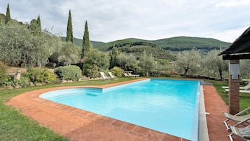 A rectangular swimming pool with a brick border, surrounded by grass and trees, with a view of hills in the background.