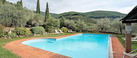 Una piscina rectangular con borde de ladrillo, rodeada de césped y árboles, con vistas a colinas al fondo.