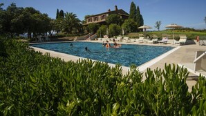 A swimming pool with lounge chairs and umbrellas, surrounded by greenery and a building in the background. - Villa Pierotta by MMega (Scarlino)