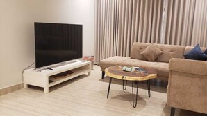 Una sala de estar con un televisor de pantalla plana sobre un mueble bajo, un sofá color beige con cojines, una mesa de centro redonda y un telón de fondo.