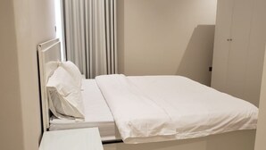 Una cama individual con ropa blanca en una habitación con pared lisa y una puerta.