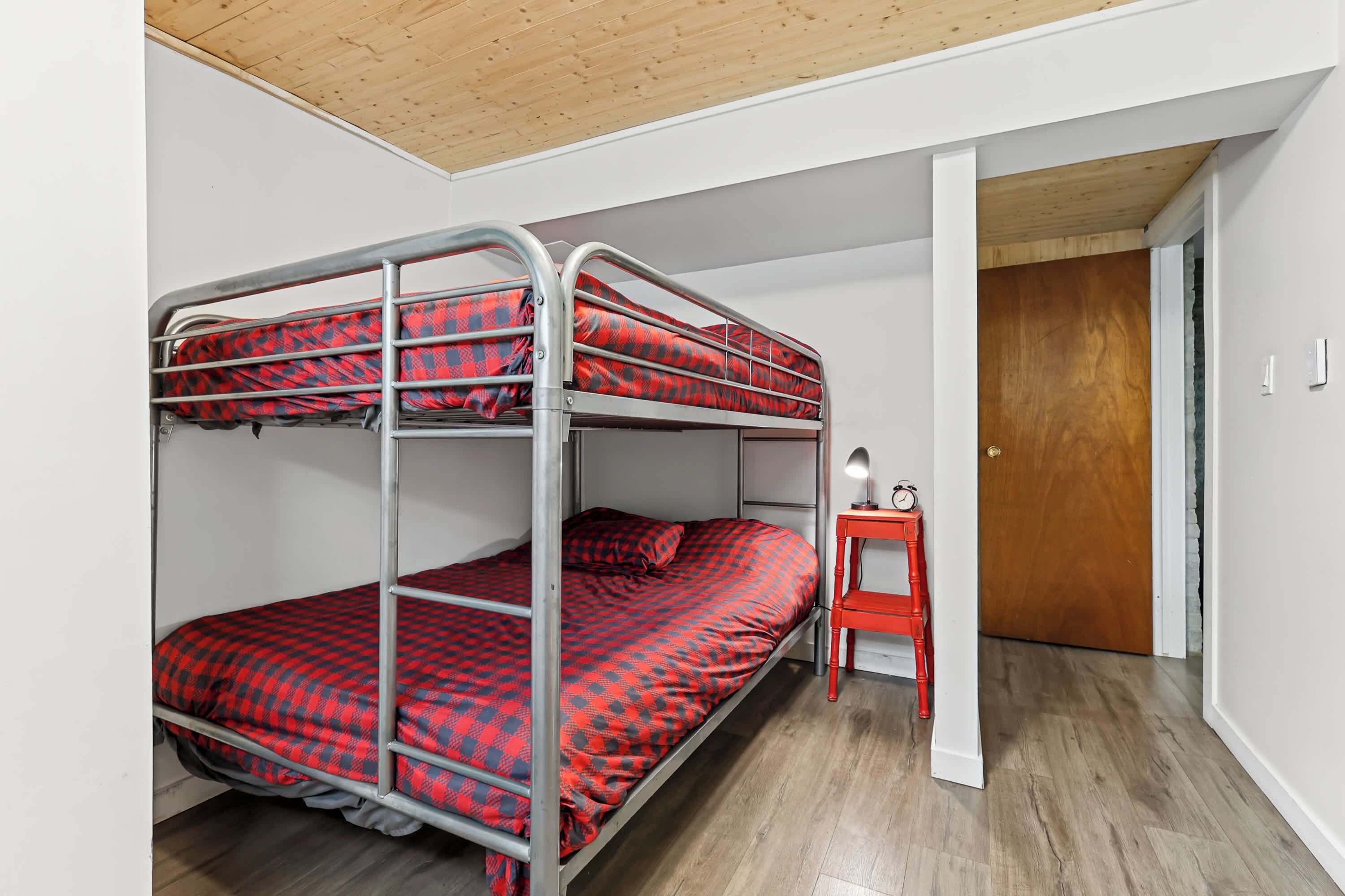 Een stapelbed met rood en zwart geruit beddengoed, een houten deur en een klein nachtkastje rood van kleur met een lamp.
