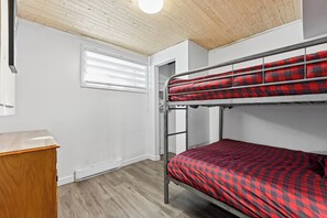 A bunk bed with a checkered blanket, a wooden desk, a window with blinds, and a wooden ceiling.