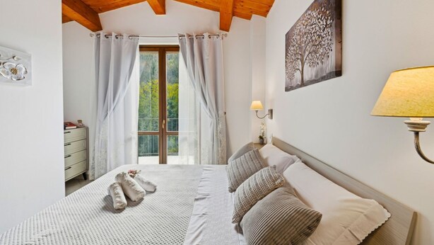 Un dormitorio con techo de madera, una cama con almohadas, una mesita de noche con lámpara, una ventana con cortinas y un cuadro en la pared.