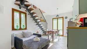 Un dormitorio con cama, ventana con cortinas, una escalera, una zona de comedor con mesa y sillas, y un espacio de cocina.