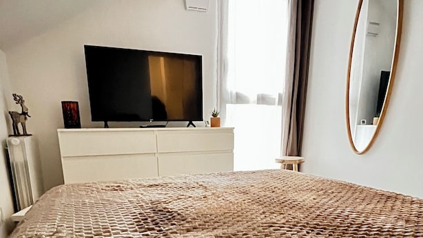 Quarto com cama grande, televisão de tela plana em cômoda branca, espelho na parede e janela com cortinas.