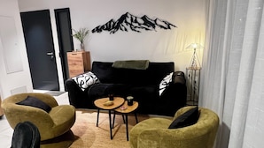 Uma sala de estar com um sofá preto, duas poltronas verdes, uma mesa de centro de madeira e um objeto decorativo na parede.