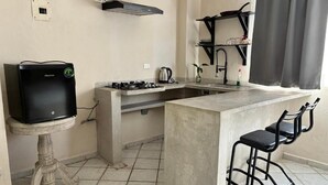 Cocina compacta con estufa a gas, un lavamanos pequeño y una barra integrada con dos sillas negras.