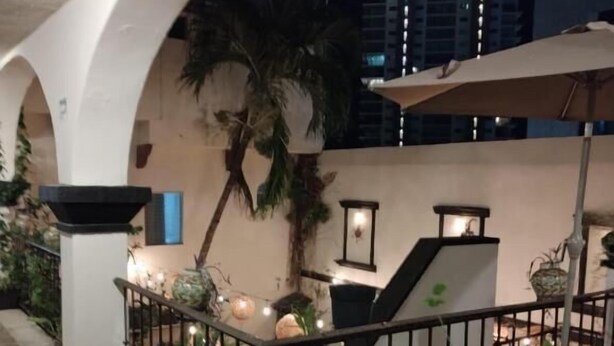 Un patio al aire libre con suelo de piedra, arcos blancos y barandilla. Hay plantas en macetas y luces colgantes.