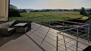 Una terraza con una mesa y sillas, baranda de metal y vistas a un campo verde y colinas distantes.
