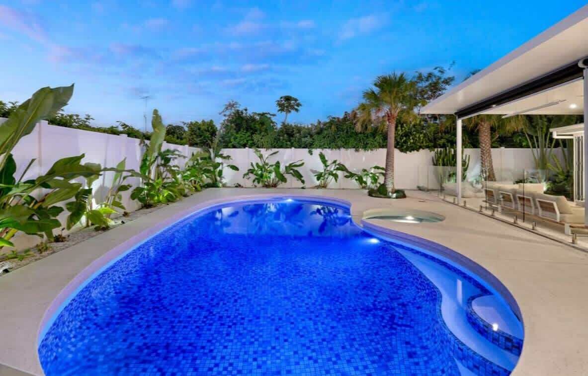 A swimming pool with a tiled edge, surrounded by a white fence and various plants.