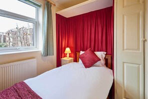 A hotel room with a bed, a nightstand with a lamp, a window with curtains, and a view of buildings outside. - Host Stay Riders Rest (Bedford)