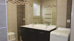 Une salle de bain avec un lavabo blanc, deux robinets, un miroir, des toilettes blanches et une poubelle.