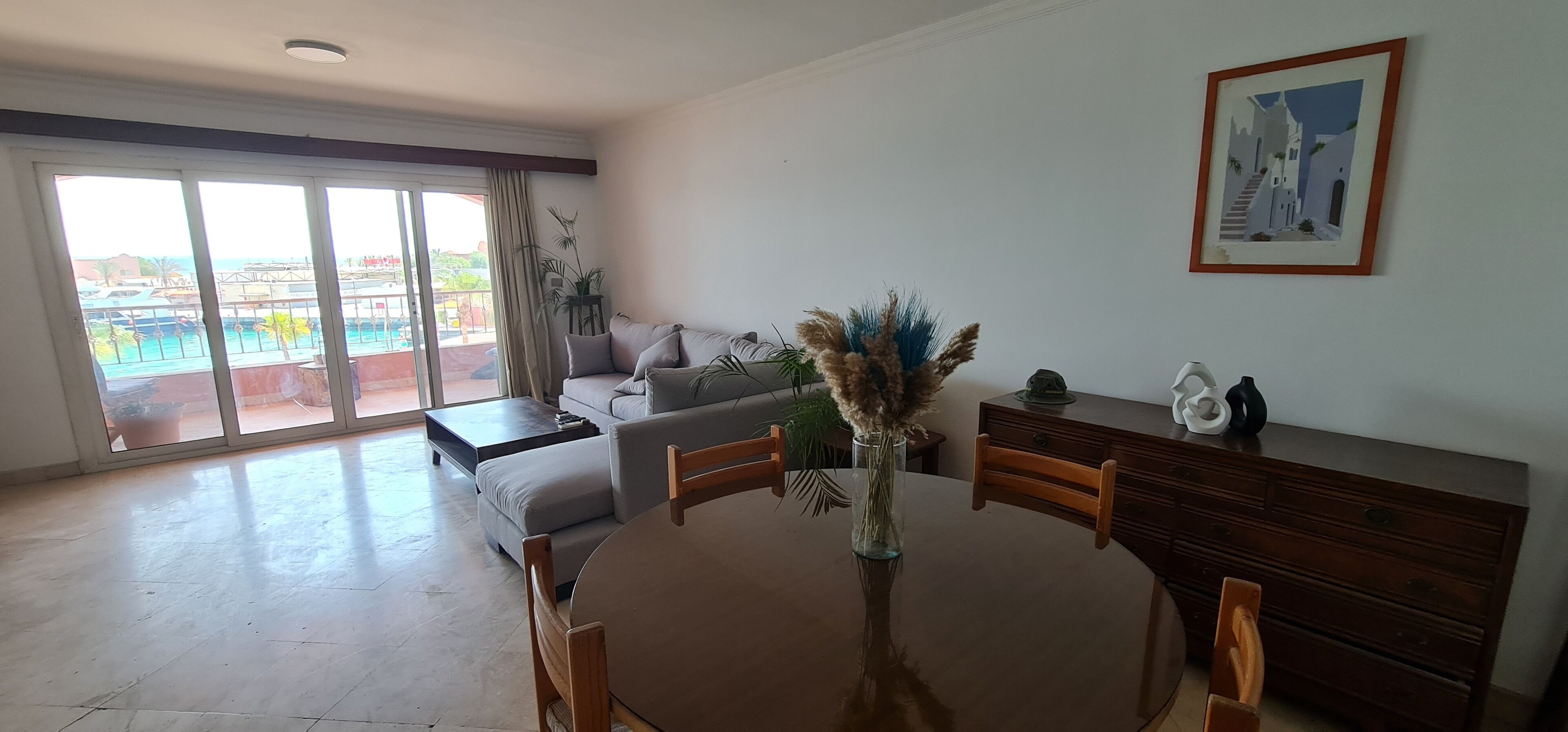 Amplia sala de estar con un gran ventanal corredero que da a un balcón, una mesa de comedor redonda con sillas, un sofá y un armario de madera.