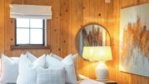 Una cama bien hecha con ropa blanca y almohadas, una lámpara de noche y un espejo grande en la pared.