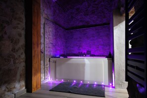 Een kamer met een stenen muur, een houten deur, een badkuip met paarse LED-verlichting en een donker vloerkleed.