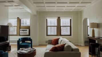 A living room with a sofa, armchairs, a coffee table, and a TV. There are large windows with blinds, a ceiling with a grid pattern, and a lamp on a table.