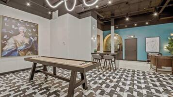 A billiards table in a modern lounge area with a geometric patterned floor, a large abstract painting, and a bar area with stools.