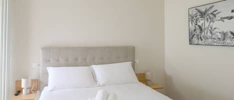 A hotel room with a bed, white linens, a headboard, a bedside table with a lamp, a wall-mounted picture, and a window with curtains.