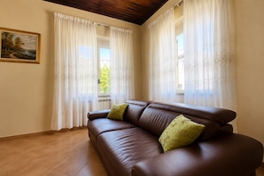 Una habitación con un sofá de cuero marrón, dos almohadas verdes, cortinas blancas con motivos florales, un cuadro de paisaje enmarcado en la pared y un techo de madera.