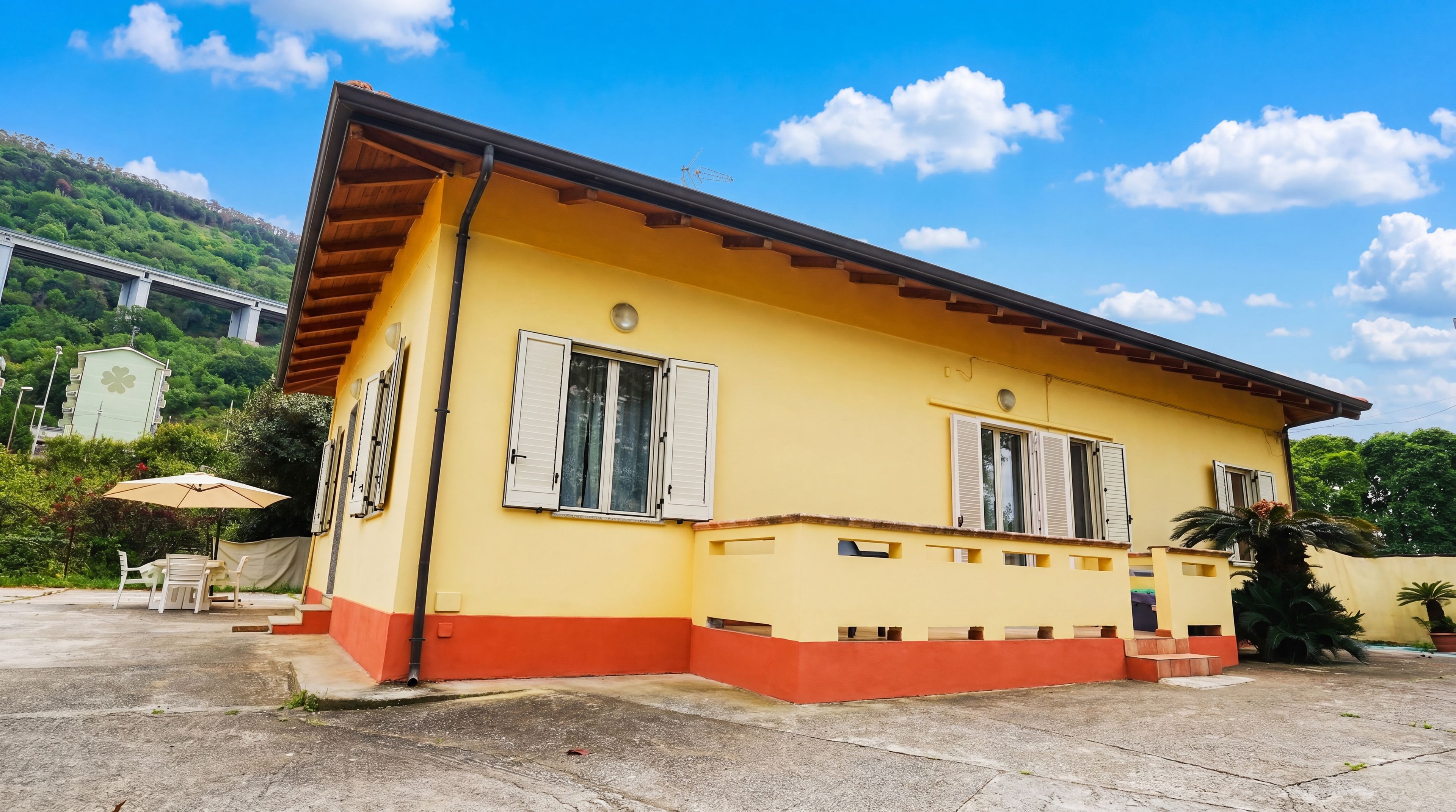 Un edificio amarillo con base roja, dos ventanas con contraventanas y un pequeño porche.