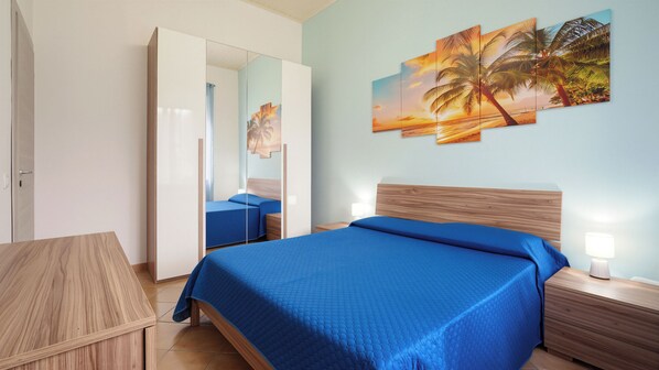 Habitación de hotel con cama, mesita de noche, espejo y un cuadro de atardecer tropical en la pared.