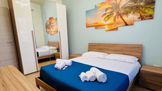 Una cama bien hecha con ropa de cama azul, un cabecero de madera y una mesita de noche con una lámpara. Un espejo grande y un cuadro con un atardecer y palmeras están en la pared.