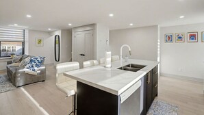 A modern kitchen with a large island, white cabinetry, and a stainless steel sink. There is a dining area with a sofa and a coffee table. The room has a large window providing natural light. - The Wynford Romantic Amalfi Escape (Elora)