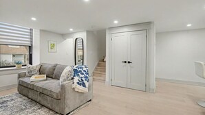 A modern living room with a grey sofa, a patterned throw blanket, and a rug. There is a white door and a staircase in the background. - The Wynford Romantic Amalfi Escape (Elora)