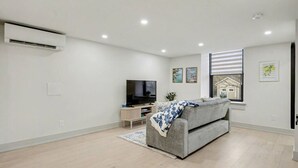 A modern living room with a grey sofa, a flat-screen TV, a small wooden side table, and a window with blinds. - The Wynford Romantic Amalfi Escape (Elora)