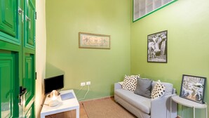 A room with a green wall, a white sofa with patterned cushions, a small white table with a monitor and a lamp, and a framed picture on the wall. - Vacation Apartment 'Guía' with Shared Terrace, Shared Garden and Wi-Fi (Tejeda)