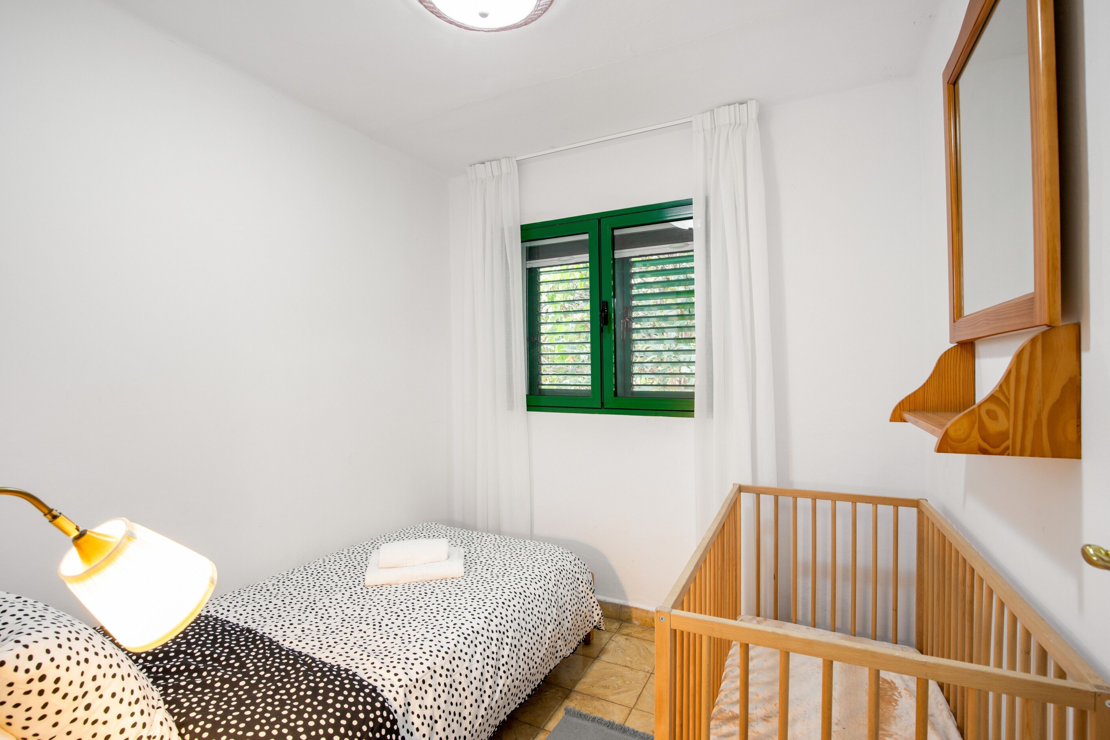 Une chambre à coucher avec un lit, un berceau, une fenêtre avec des volets verts et un miroir fixé au mur.