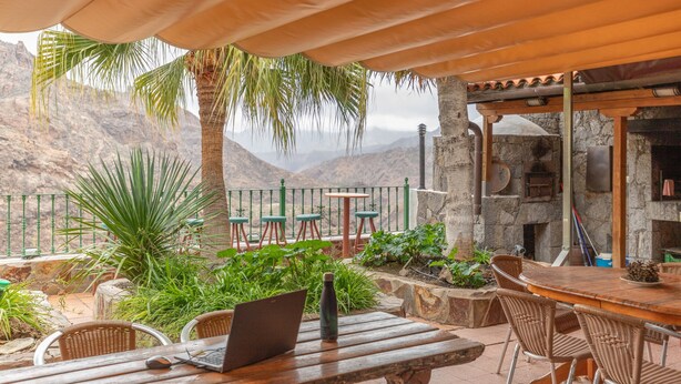 An outdoor patio with a wooden table, wicker chairs, and a laptop.
