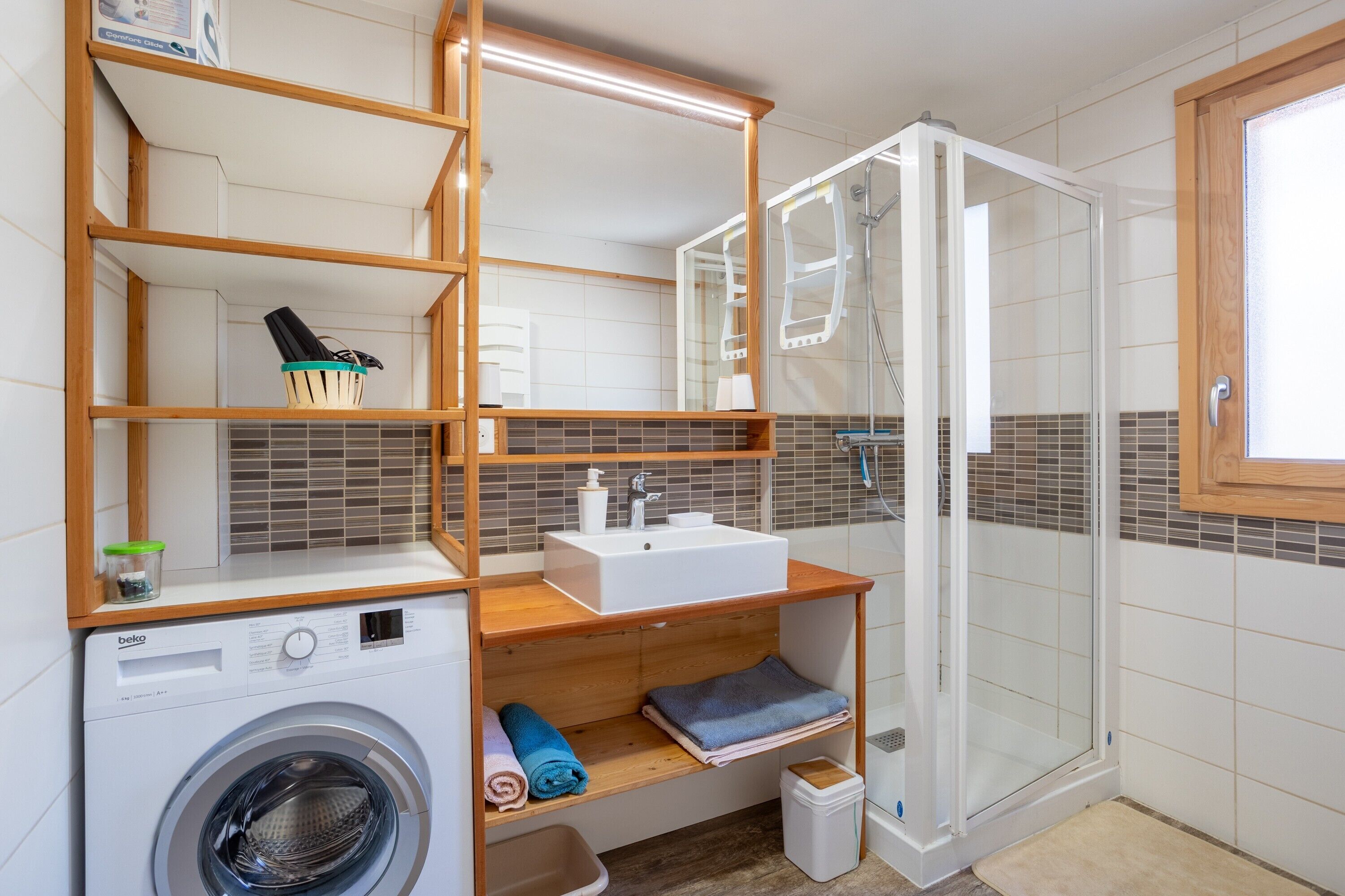 A compact bathroom with a washing machine, a sink, a glass shower enclosure, and built-in shelves.