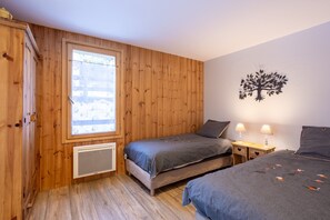 Ein Zimmer mit Holzvertäfelung, einem Fenster, zwei Betten mit grauer Bettwäsche, einer an der Wand befestigten Baumdekoration und einem kleinen Nachttisch mit Lampen.