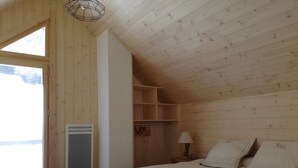 Une chambre avec un plafond en bois, un lit recouvert de linge de lit blanc, une table de chevet avec une lampe et une fenêtre donnant sur l’extérieur.