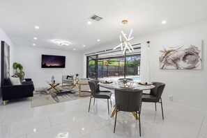 A modern living room with a round dining table, grey chairs, a black sofa, and a large window with a view of greenery. - Remodeled, Close to Beach, With Heated Pool (Naples)
