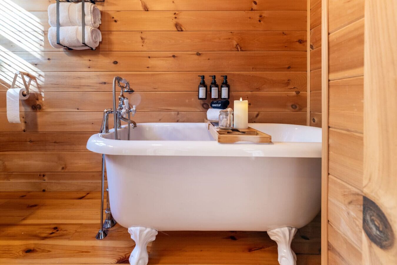 Une salle de bain rustique, avec une cabine en bois, une baignoire sur pieds, une étagère en bois où sont rangés les articles de toilette et un porte-serviettes.