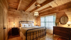 Un dormitorio de cabaña de madera con cama, mesitas de noche, cómoda, candelabro y ventana con cortinas.