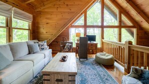 Interior acogedor de una cabaña con techo de madera, ventanas amplias, sofá, escritorio y computadora.